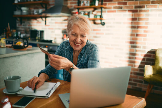 Happy senior woman paying online with credit card in home kitchen