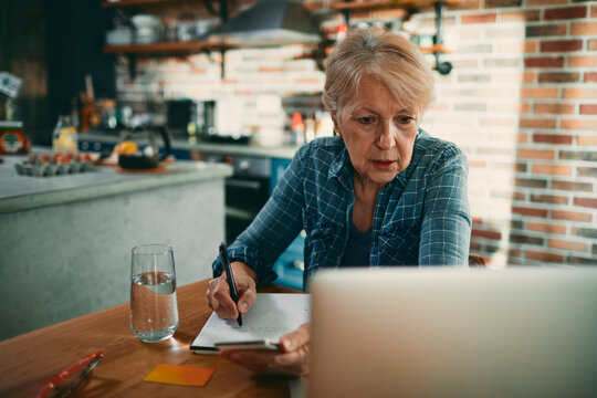Senior woman writing notes while working on laptop at home kitchen