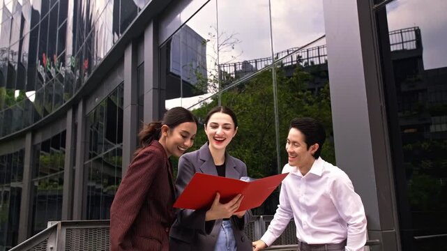 Three professionals review documents in a red folder outside a modern glass office, smiling and discussing plans.