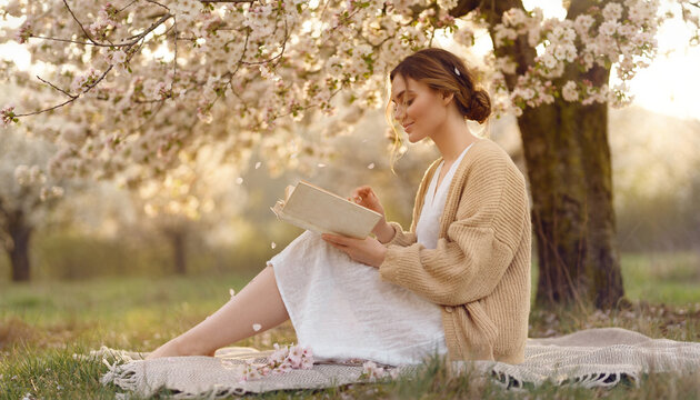 Young woman reading book in spring blossom orchard at sunset