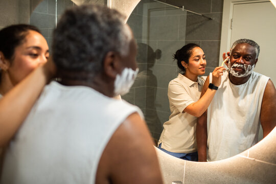 Caregiver helping senior man shave at home bathroom mirror