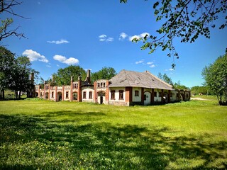 Obraz premium An abandoned historic palace with red brick and plaster walls stands in a grassy field under a clear blue sky. The building displays a series of arches and large, arched windows
