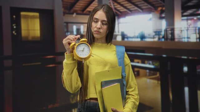 Young woman student holding yellow alarm clock and a stack of folders in a school building; anxious deadline pressure.