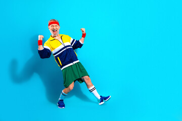 Obraz premium Energetic senior man in retro workout outfit celebrating fitness and victory, cheering with clenched fists against a bright blue background