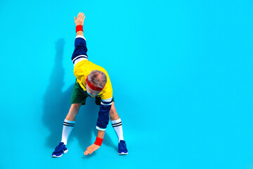 Obraz premium Senior man stretching in retro sportswear on a bright blue background showing active lifestyle, fitness, balance and healthy aging with colorful headband and wristbands