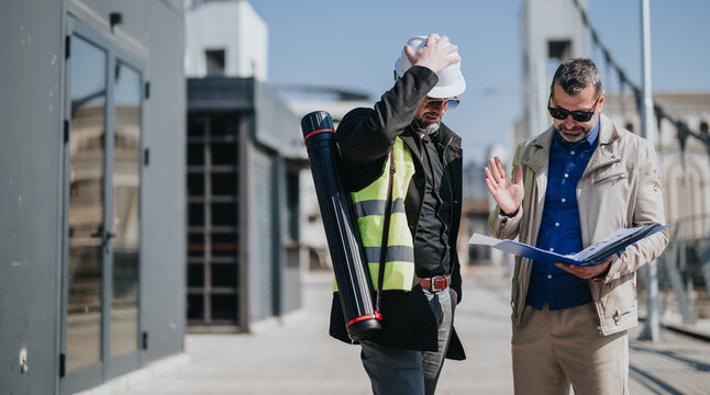 Two professionals review building plans on an outdoor construction site, one wearing a hard hat. They discuss documents and drawings while standing near a modern building under construction.