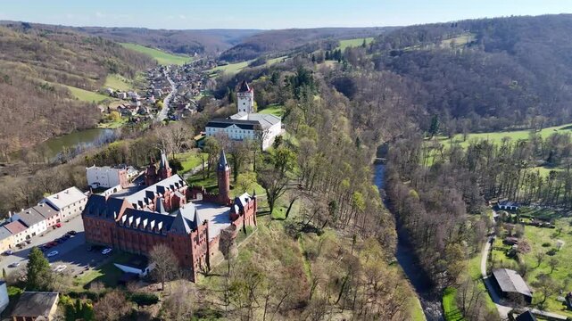 Rightward movement as drone slowly rotates and descends reveals Hradec nad Moravic&iacute; castle complex in Czech Republic with river valley, trees and rolling countryside landscape.