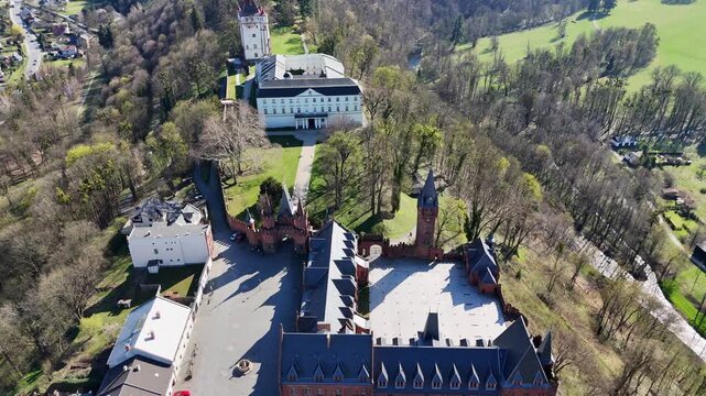 Backward flight as drone slowly tilts camera upward reveals Hradec nad Moravic&iacute; castle complex in Czech Republic with surrounding hills, village and wide countryside landscape.
