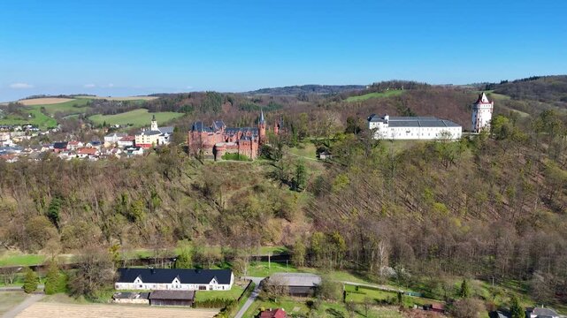 Drone flying backward reveals Hradec nad Moravic&iacute; castle on hill with surrounding countryside, village and rolling landscape under clear blue sky.