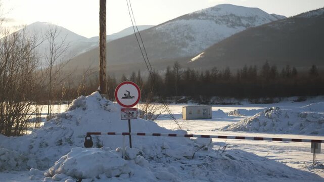 A snow-covered area shows a warning sign near a frozen lake. Mountains rise in the backdrop. The scene captures the cold winter day, showcasing nature and safety measures.