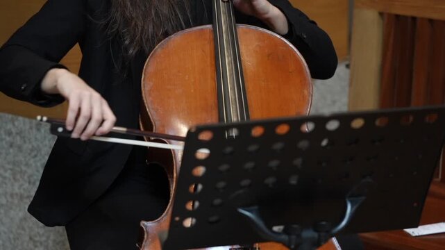 Person playing cello with hands on strings and fingerboard while reading sheet music in indoor performance setting