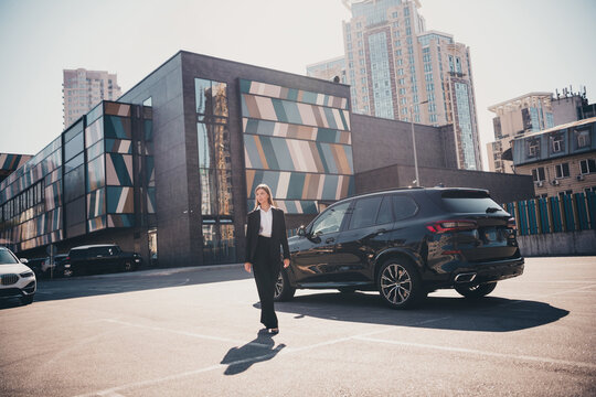 Businesswoman walking past luxury SUV in modern urban parking lot by contemporary office buildings