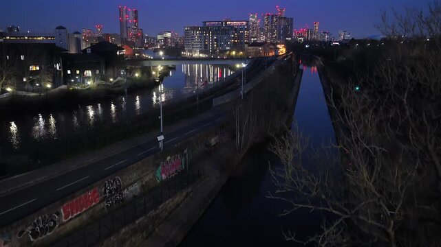 Aerial video at night showing the River Irwell, Bridgewater Canal, and Metrolink tracks leading to the Manchester skyscraper skyline, UK.