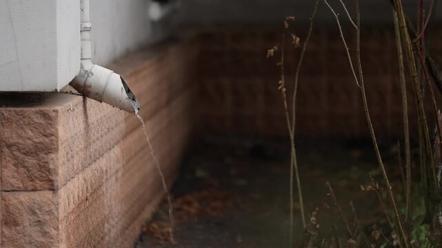 Rainwater flowing from house drain pipe in close-up. Water stream pouring from roof gutter downspout near building wall, outdoor drainage system detail during rainy weather with blurred background