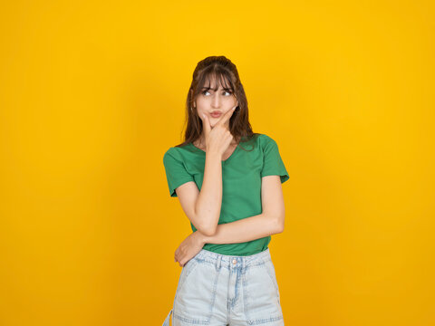 Thoughtful caucasian woman touching chin and looking aside with thinking expression, considering decision concept, wearing green t shirt and denim, isolated yellow studio background with copy space.