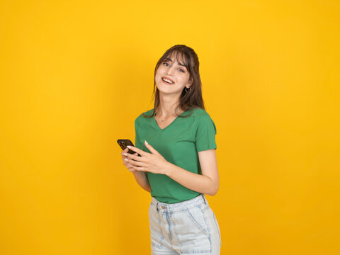 Smiling caucasian woman holding smartphone with relaxed cheerful expression, wearing green t shirt and denim, isolated on yellow studio background with copy space. Communication lifestyle concept.