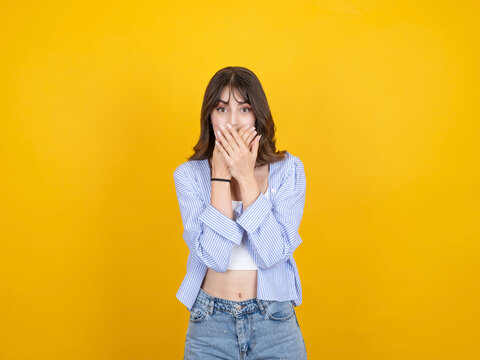 Shocked caucasian woman covering mouth with both hands, reacting with surprise concept idea image, wearing striped shirt and denim, isolated on bright yellow studio background with copy space.