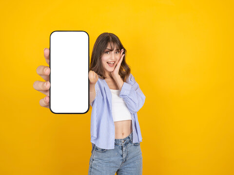 Excited caucasian woman showing smartphone with blank screen mock up, holding cheek with joyful expression, wearing striped shirt and denim, isolated on yellow studio background with copy space.