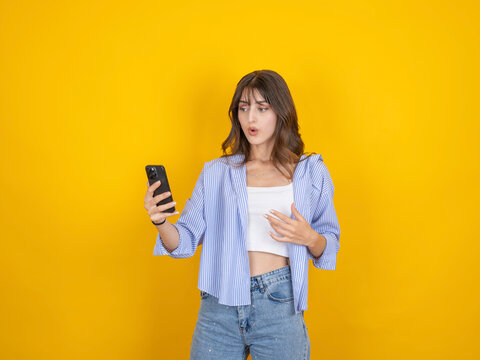 Surprised caucasian woman looking at smartphone with shocked expression, reacting to message, wearing striped shirt and denim, isolated on bright yellow studio background with copy space.