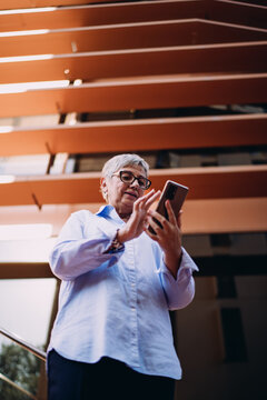 Elderly woman stands under modern structure, focused on smartphone while typing. Calm face reflects confidence with tech in urban environment.
