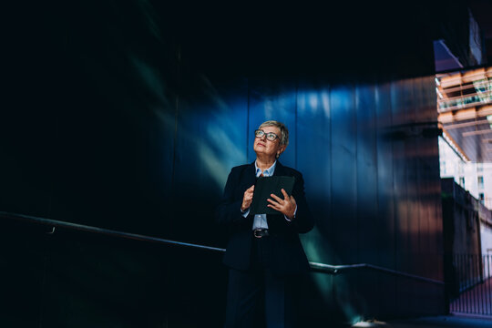 Focused mature businesswoman holding tablet while looking upward. Scene symbolizes digital strategy, future thinking, and decision-making in mobile-driven industries.