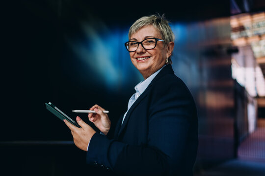 Senior female professional smiling while working on tablet with stylus. Represents mobile flexibility, digital command, and joyful engagement in smart work tools.