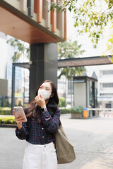 Fototapeta premium Young woman confidently walking in an urban environment while wearing a face mask for protection and holding a modern smartphone, representing changing city lifestyles