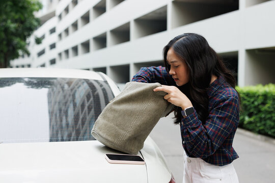 Young Asian woman meticulously searching through her handbag for missing items or car keys while standing next to her vehicle in a parking lot, reflecting a moment of frustration