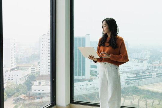 Asian professional woman standing by a large office window, holding a laptop and contemplating the urban city view, representing concentration, future vision, and contemplation during work