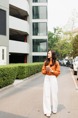 Fototapeta premium Young Asian woman on a daily walk through an urban street, confidently holding her smartphone while looking away, with modern buildings and green hedges in the background
