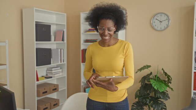 Woman holding a clipboard flips pages and adjusts glasses in a studio office with bookshelf, clock and houseplant visible; contemplation.