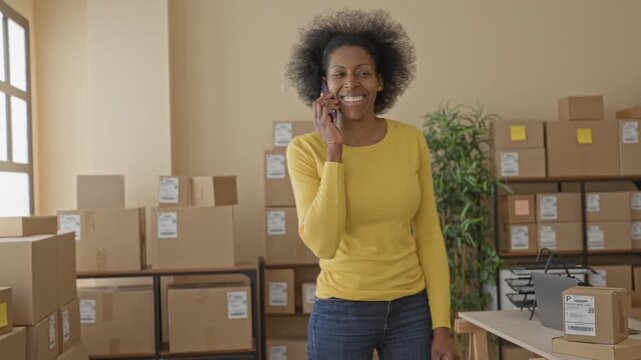 Woman talking on phone holding parcel and gesturing with hand in packing area inside building; small business cheerful.
