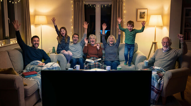 A multigenerational family gathers in a cozy living room, immersed in a moment of joyful unity. Cheering and celebrating as they watch television together, highlighting the comfort and connection.