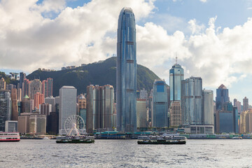 Fototapeta premium Hong Kong skyline dominated by the International Finance Centre, ferries crossing Victoria Harbour and a bustling waterfront of high-rise buildings against green hills and dramatic clouds