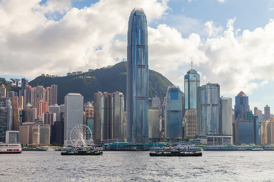 Hong Kong skyline dominated by the International Finance Centre, ferries crossing Victoria Harbour and a bustling waterfront of high-rise buildings against green hills and dramatic clouds