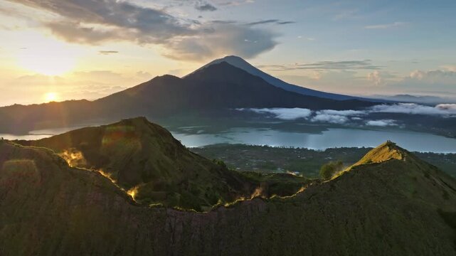 Bright sunset over Mount Batur volcano caldera revealing smoking vents, Lake Batur and the distant Mount Agung, capturing Bali dramatic volcanic evening landscape. Aerial view drone flight panorama