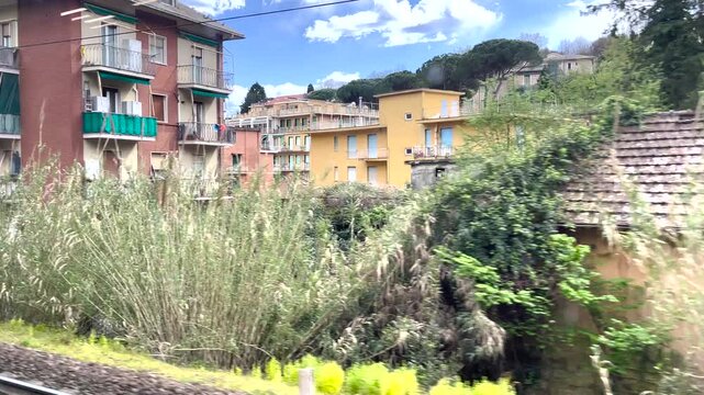 Italian regional train window view passing Sestri Levante residential area. Sunny day journey through Ligurian coast showing traditional colorful apartments and Mediterranean greenery. Authentic
