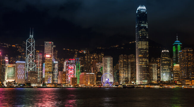 A panoramic night view of Hong Kong skyline across Victoria Harbour, featuring illuminated skyscrapers, colorful waterfront lights and reflections on the water