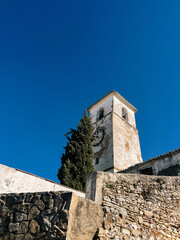 Old white church tower behind a stone wall under a clear blue sky. Colmenar, Malaga, Andalusia, Spain.