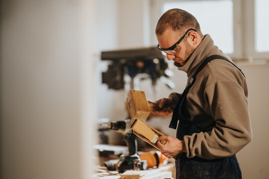 A carpenter wearing safety glasses planes a wooden block with a mallet and hand plane. The craftsman works carefully at a bench in a well-lit workshop.