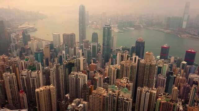 Aerial view of Hong Kong&rsquo;s dense urban skyline, with Central and Wan Chai skyscrapers rising above Victoria Harbour in hazy daylight, showing modern architecture and waterfront city life.