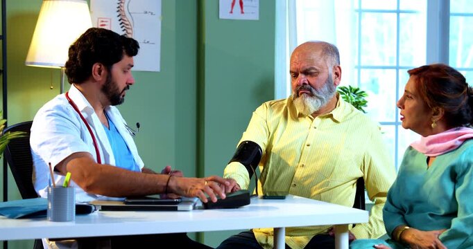 Professional Indian young male doctor checking blood pressure of senior retired man across a table in a modern clinic and showing dissatisfaction with results; healthcare and old age concept.