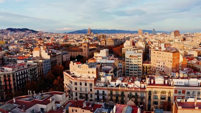 Aerial cityscape of Barcelona, Spain at golden hour, featuring dense rooftops, the Sagrada Familia and Torre Glories on the skyline with mountains in the background.