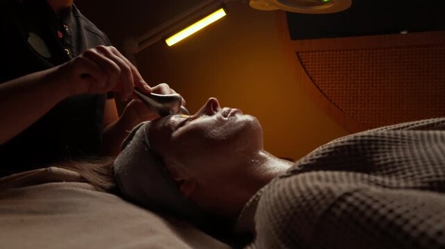 Relaxing facial spa treatment with esthetician gently massaging client skin using stone tools under warm ambient lighting in a tranquil wellness setting