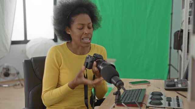 Woman adjusting camera with hand at desk in studio beside microphone and green screen, wearing yellow sweater; concentration creativity.