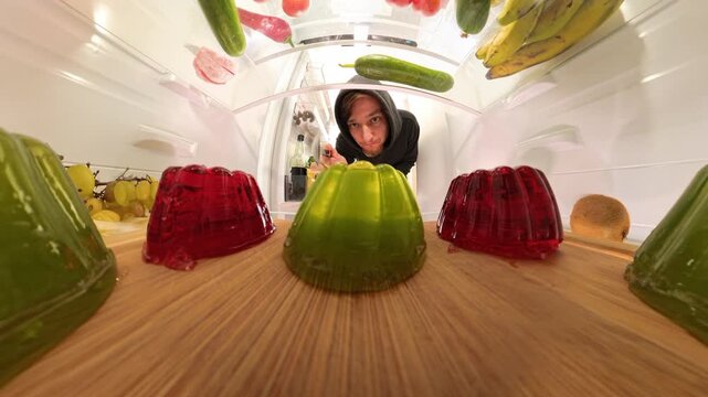 Dolly zoom view from inside the refrigerator as a man uses a spoon to taste green jelly, capturing the wobble and soft texture of the dessert in a close-up shot