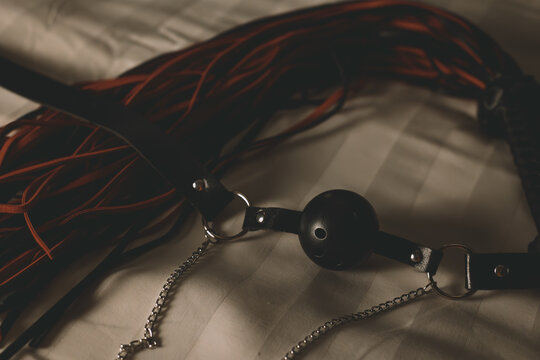 Ball gag and leather flogger resting on wrinkled bed sheets