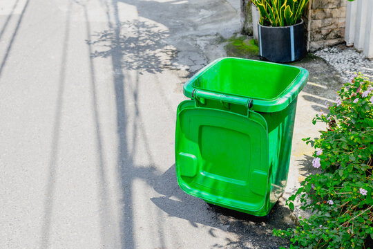 Green trash bin standing open on sunlit sidewalk near residential wall