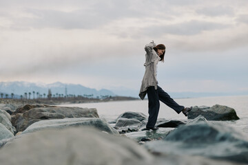 Obraz premium A young woman balances carefully on rocks by the sea under a cloudy sky. Casual outfit and natural coastal landscape create a peaceful and adventurous mood.