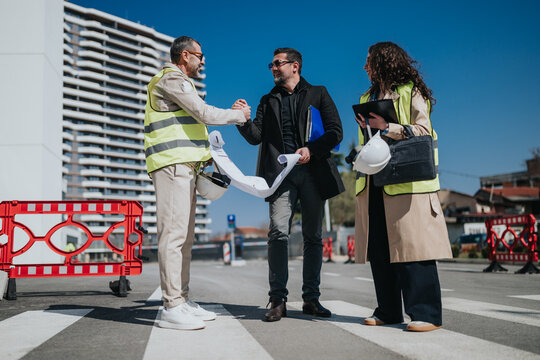 Three construction professionals review blueprints and shake hands on an urban roadwork site. A client and site managers discuss plans beside safety barriers and helmets under a clear sky.